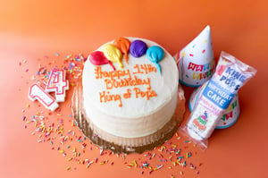 A birthday cake against an orange background surrounded by sprinkles, party hats, birthday candles, and a Birthday Cake Pop